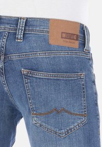 Jeans de mezclilla azul con una etiqueta de cuero en la parte trasera que dice "MUSTANG True Denim." Presenta detalles de costura y un único bolsillo trasero.