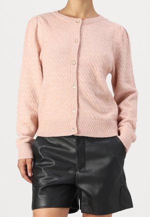 Vrouw die een lichtroze, getextureerde, met knopen gesloten cardigan draagt en zwarte leren shorts, staand met één hand in de zak.