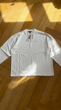 Camisa branca de manga longa com decote redondo, feita de tecido suave, apresentando uma etiqueta com texto. Disposição plana sobre um chão de madeira.