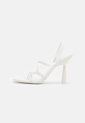 Sandale blanche à talons hauts avec des fines lanières, une bride arrière et un talon bloc évasé sur un fond clair uni.