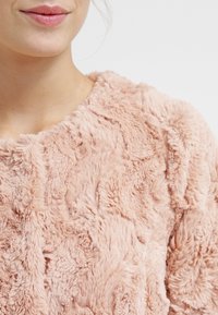 Sweter z sztucznego futra w delikatnym różu, charakteryzujący się teksturowaną powierzchnią o różnej długości i wzorach. Okrągły dekolt z subtelnym detalem szycia.