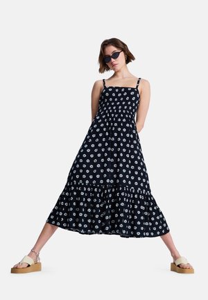 Robe noire sans manches avec imprimés floraux blancs, corsage froncé, ourlet à volants et bretelles réglables, associée à des sandales compensées beiges.