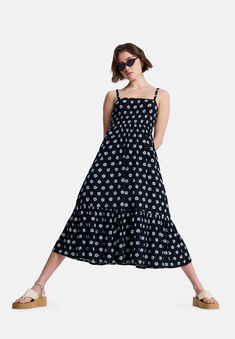 Μαύρο αμάνικο φόρεμα με λευκά λουλουδάτα σχέδια, σούρα στο μπούστο, βολάν στο τελείωμα και ρυθμιζόμενους ιμάντες, συνδυασμένο με μπεζ σανδάλια πλατφόρμα.