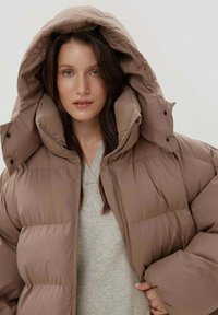 Brązowa kurtka puchowa z dużym kapturem, o pikowanym wzorze i gładkiej fakturze. Noszona na jasnoszarym swetrze, zapinana na zatrzaski.