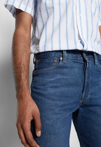 Vaqueros de mezclilla en azul oscuro con una textura desgastada, con un diseño clásico de cinco bolsillos y un botón de cobre en la cinturilla. Llevados con una camisa a rayas.