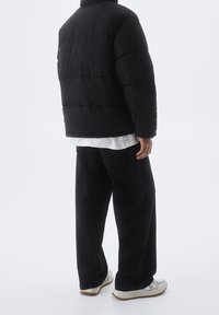 Doudoune noire avec col haut, design matelassé et rembourrage épais. Portée par-dessus une chemise blanche et associée à un pantalon large noir et des baskets.