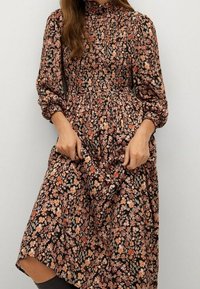 Robe longue à fleurs avec un fond noir orné de petites fleurs orange et crème, taille froncée, col montant et manches bouffantes.