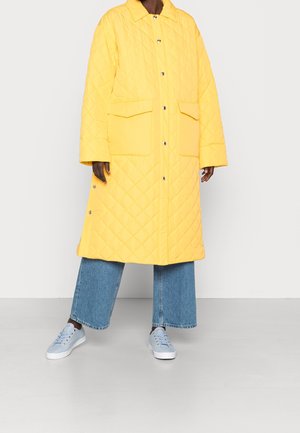 Personne portant un long manteau matelassé jaune avec de grandes poches avant, un jean bleu large et des baskets bleu clair, debout sur un sol gris.