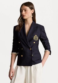Blazer bleu marine à double boutonnage en tissu lisse, avec des boutons dorés et un blason brodé sur la poitrine. Manches retournées avec des bordures blanches.