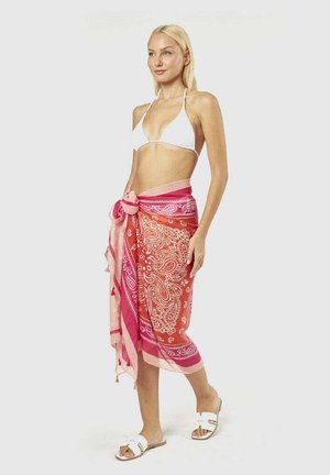 Donna bionda con top bikini bianco, sarong rosso e rosa a fantasia paisley annodato in vita e sandali bianchi slip-on, in piedi su sfondo bianco.