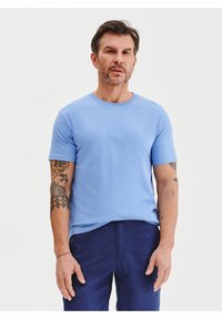 Jasnoniebieska bawełniana koszulka z krótkim rękawem i okrągłym dekoltem, charakteryzująca się gładką fakturą i minimalistycznym stylem. Noszona z ciemnoniebieskimi spodniami.