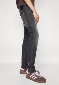 Fekete farmernadrág elhasználódott felülettel, slim fit szabású, elöl és hátul zsebekkel, bordó és fehér sportcipőkkel párosítva.