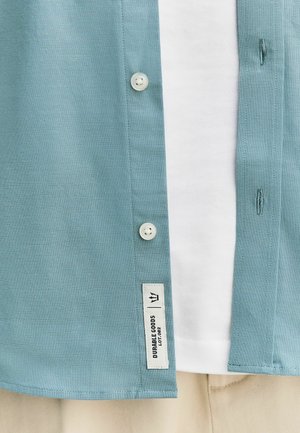 Hellblaues Hemd über einem weißen T-Shirt und beigen Hosen, mit einem „Durable Goods“-Label nahe dem unteren Saum.