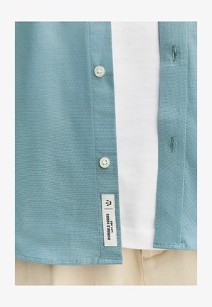 Cămașă cu nasturi albastru deschis peste tricou alb și pantaloni bej, cu etichetă „Durable Goods” lângă marginea de jos.