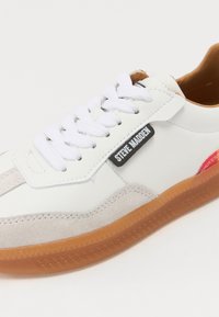 Valkoiset sneakerit, joissa on nahka päällinen, mokkanahka yksityiskohtia ja kumipohja. Varustettu litteillä valkoisilla nauhoilla ja mustalla logo-tarralla sivulla.
