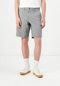 Personne debout portant un short gris jusqu'aux genoux, des chaussettes blanches, et des baskets crème avec des semelles en gomme sur un fond blanc.