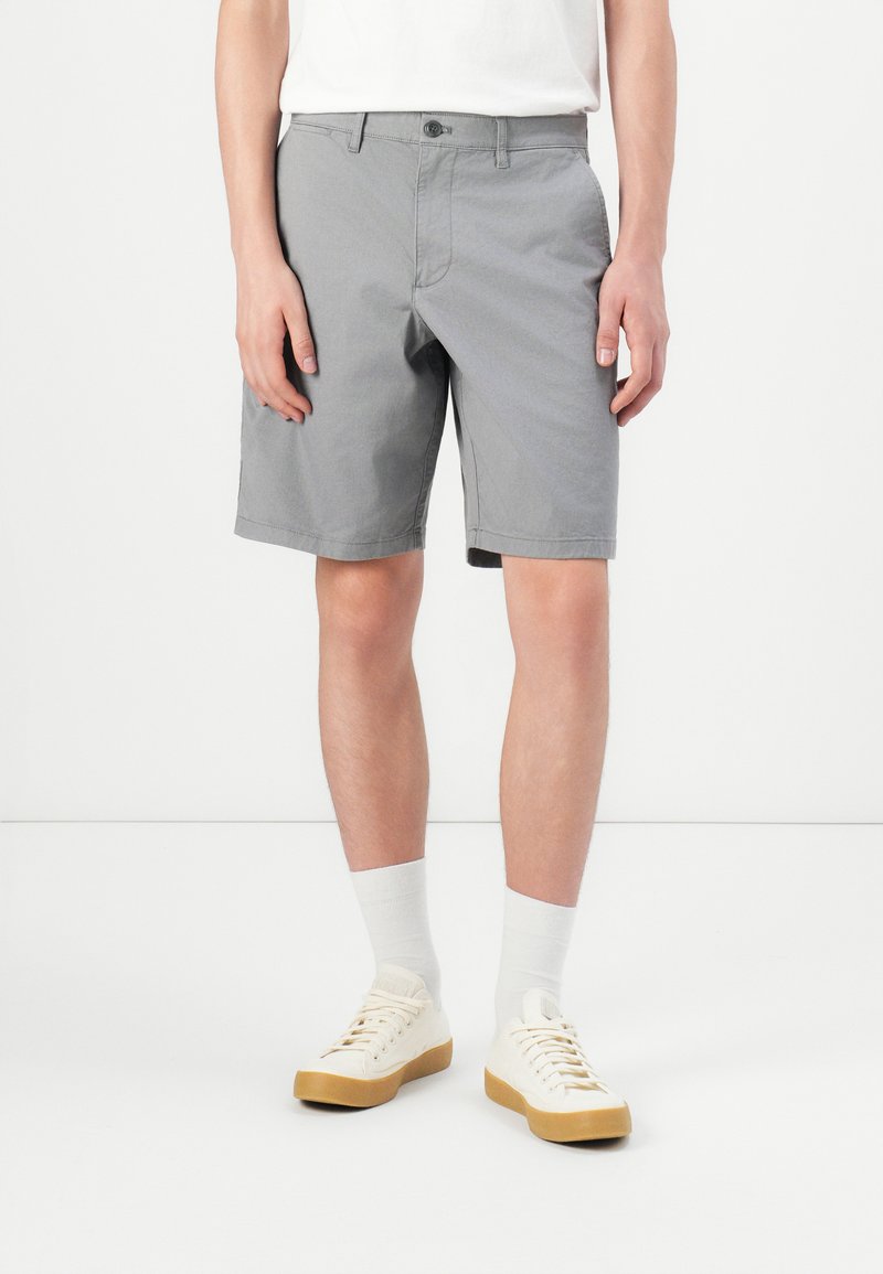 Personne debout portant un short gris jusqu'aux genoux, des chaussettes blanches, et des baskets crème avec des semelles en gomme sur un fond blanc.