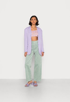 Blazer oversized em tom lilás, top curto floral, calças de corduroy largas em verde e sandálias de salto lavanda sobre um fundo neutro.