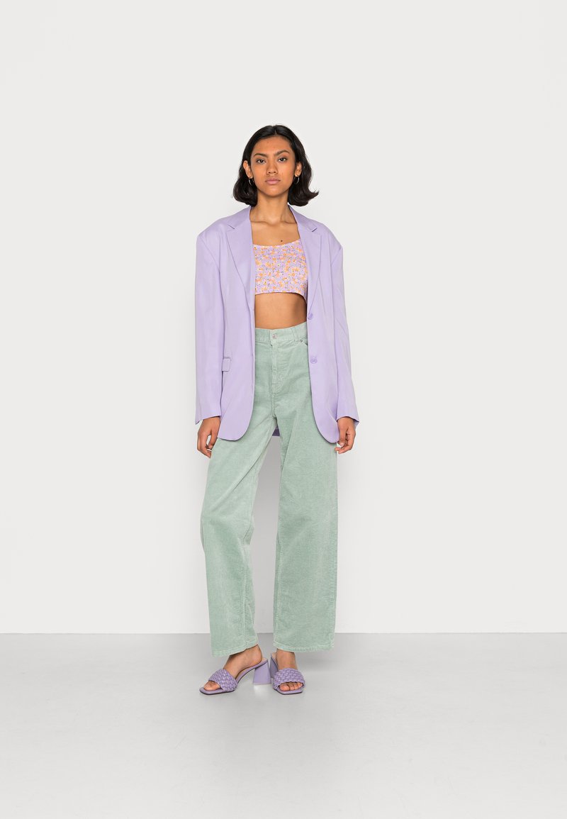 Blazer oversized em tom lilás, top curto floral, calças de corduroy largas em verde e sandálias de salto lavanda sobre um fundo neutro.