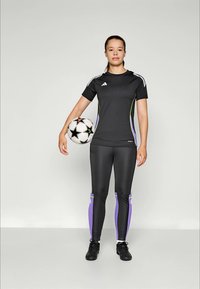 Ung kvinne i svart og lilla treningstøy som holder en hvit og svart fotball mot en enkel lys bakgrunn.