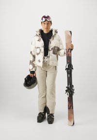 Daunenjacke mit weißen und braunen Mustern, schwarzes Basisteil, beige Skihose, hält schwarze Skier und einen Helm mit rosa getönten Skibrille.