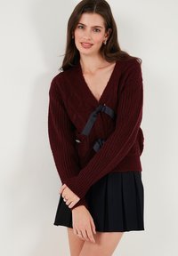 Burgundy cable-knit cardigan with a deep V-neck, featuring a black ribbon tie at the waist. Long sleeves and textured fabric.