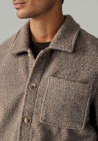 Jachetă din fleece texturat, de culoare maro, cu guler, două nasturi și un buzunar aplicat pe partea stângă a pieptului. Material moale și călduros.