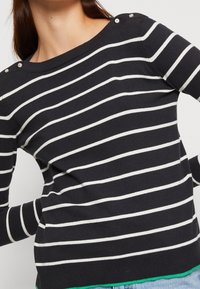 Navyblauer und weißer gestreifter Pullover mit Bootsausschnitt, dekorativen Knöpfen an den Schultern und einem grünen Akzent am Saum.