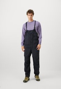 Black, durable overalls with adjustable straps and a front pocket, worn over a light purple long-sleeve shirt; styled with rugged shoes.