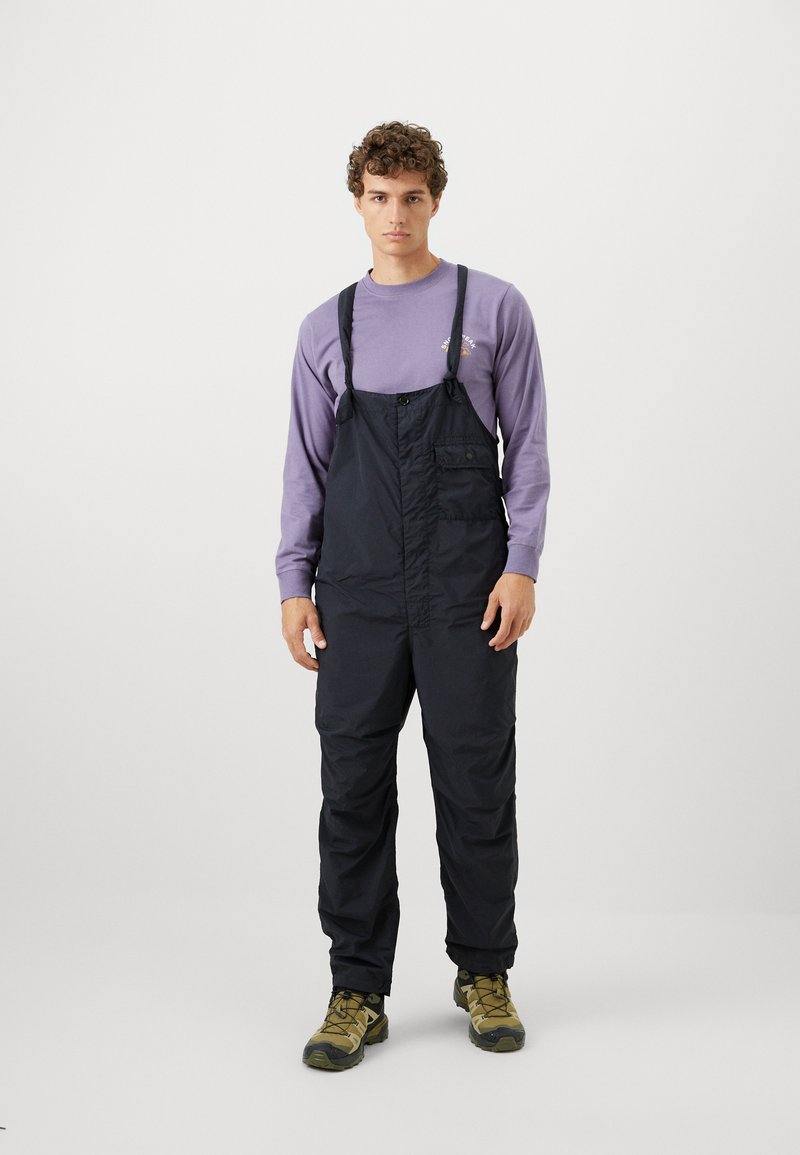 Black, durable overalls with adjustable straps and a front pocket, worn over a light purple long-sleeve shirt; styled with rugged shoes.