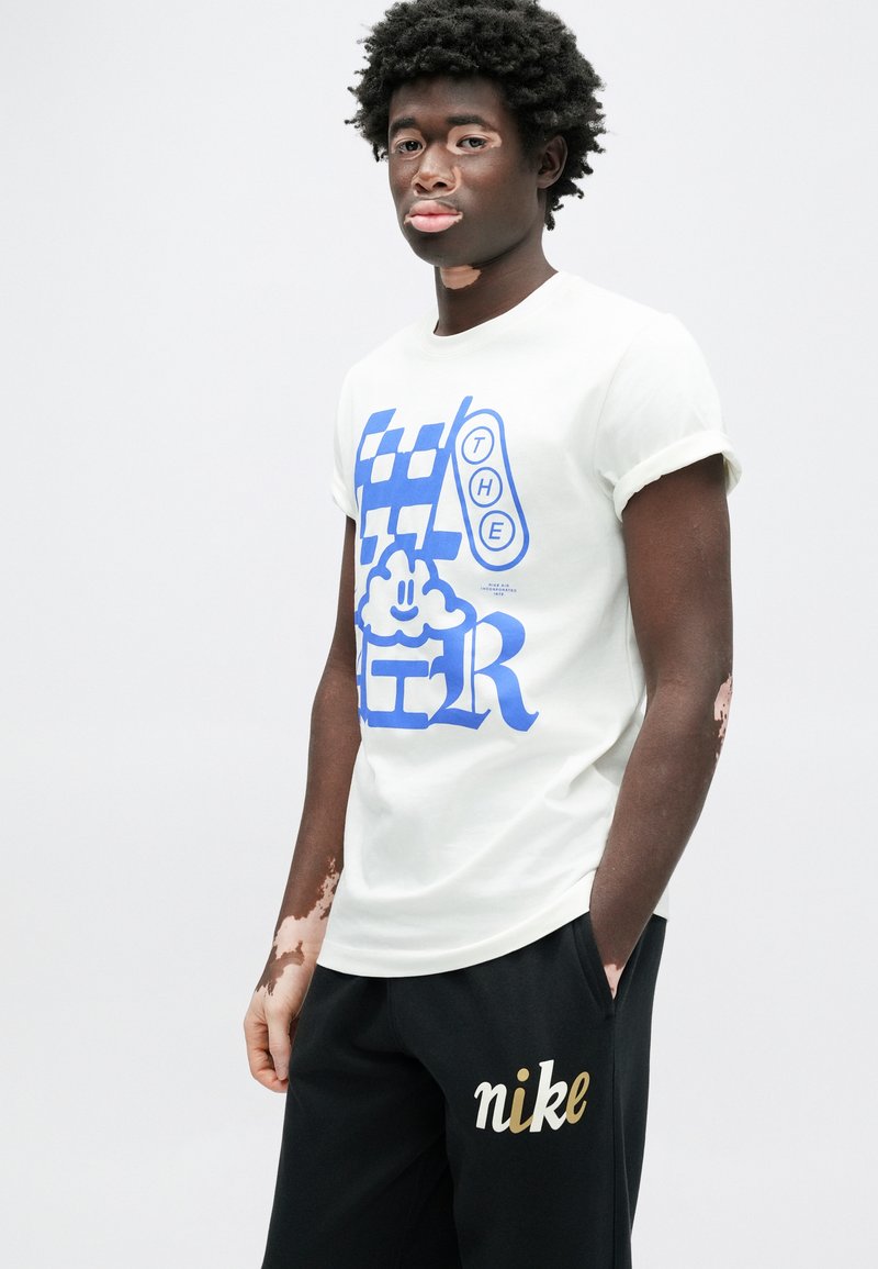T-shirt en coton blanc avec un texte graphique bleu audacieux, associée à un pantalon de survêtement noir avec le logo "nike" doré. Coupe décontractée avec manches retroussées.