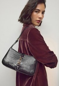 Sac à main noir embossé croco avec une forme incurvée, doté d'un accent en chaîne dorée et d'une bandoulière amovible. La surface texturée renforce les détails.