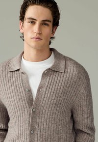 Cardigan gri cu nervuri, realizat din lână moale, cu închidere cu nasturi, guler și purtat peste un tricou alb. Textură netedă.