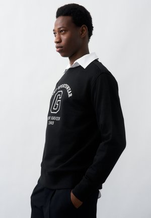 Ung mand i sort sweatshirt med hvid tekst og hvid skjorte med krave står sidelæns med hænderne i lommerne foran en ensfarvet baggrund.