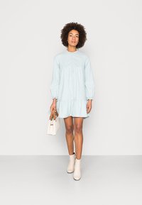 Vestido listrado azul claro com bainha rodada, mangas longas e detalhes em botões. Combinado com botas de tornozelo bege e uma mala branca.