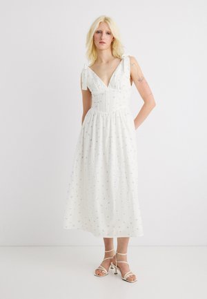 Mujer rubia con tatuaje en el brazo que lleva un vestido midi blanco con pequeño estampado floral azul y tirantes atados en los hombros, de pie con tacones blancos con tiras.