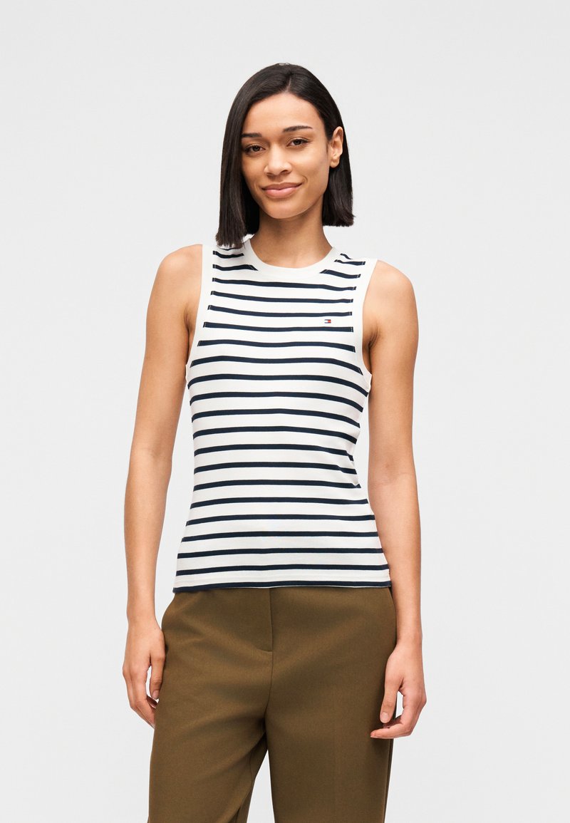 Λευκό αμάνικο τοπ με ναυτικές οριζόντιες ρίγες, στρογγυλή λαιμόκοψη και λογότυπο κοντά στο στήθος· συνδυασμένο με καφέ παντελόνι φαρδιάς γραμμής.