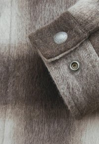 Brown and cream plaid wool fabric features a thick, textured weave. Fastened with a metal snap and grommet detail on the cuff.