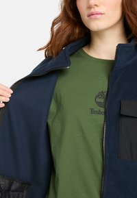 Granatowa polarowa kurtka z czarnymi akcentami, zamek błyskawiczny, boczna kieszeń, noszona na zielonym T-shircie z logo drzewa. Widoczny teksturowany materiał.