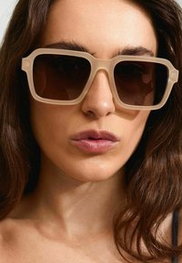 Gafas de sol grandes, translúcidas en color beige con lentes oscuros; diseño de montura cuadrada; estilo minimalista con bordes marcados.