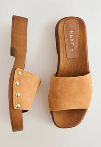 Sabots en suède beige clair avec une large bride et des clous en laiton, posés sur une semelle en bois marron à plateforme avec une finition texturée.