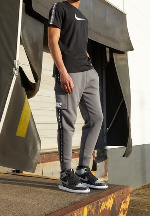 Mann trägt schwarzes Nike-T-Shirt, graue Nike-Sweathosen mit Seitenstreifen und schwarze und graue Nike High-Top-Sneaker, steht in der Nähe einer Lagertür.