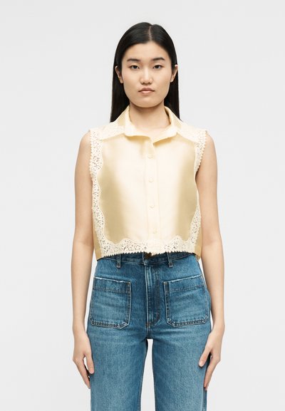 Γυναίκα που φοράει αμάνικο κρεμ σατέν μπλουζάκι με δαντέλα στο τελείωμα και μπλε ψηλόμεσο τζιν με εμπρόσθιες θήκες, στέκεται μπροστά σε απλό φόντο.