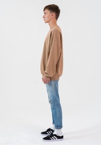 Brązowa, oversize'owa bluza z raglanowymi rękawami, zestawiona z przetartymi jasnoniebieskimi dżinsami i czarnymi sneakersami z białymi akcentami.