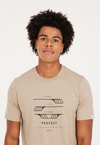 Camiseta de algodón color beige con texto gráfico y líneas en negro. Manga corta, cuello redondo, con un sutil logo en la manga.