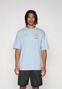 T-shirt en coton bleu clair à manches courtes, arborant un logo NY orange, établi en 1993. Porté avec un short de sport noir perforé.