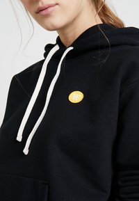 Svart huvtröja i bomull med vita dragsnören och en liten gul cirkulär logotyp med ett "W" broderat på bröstet.