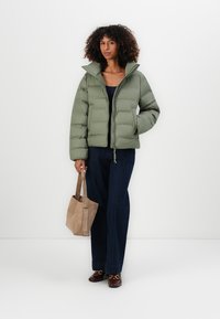 Olijfgroene pufferjack met een hoge kraag, ritssluiting aan de voorkant en een elastische rand, gecombineerd met donkere denimbroeken en een lichtbruine totebag.