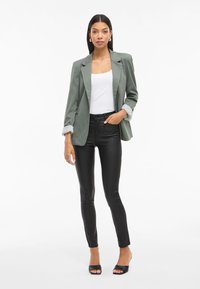 Grijze blazer met opgerolde mouwen, witte tanktop, zwarte leren broek en zwarte open-toe hakken, staand tegen een witte achtergrond.