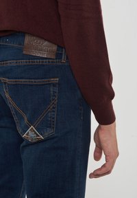 Lato destro di una persona che indossa jeans di denim blu scuro e un maglione rosso scuro, mostrando la tasca posteriore dei jeans e il tag in pelle del marchio.